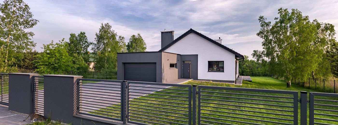 Modernes Einfamilienhaus mit weißer Fassade, dunklem Dach und großem Metallzaun im Vordergrund, umgeben von Bäumen.